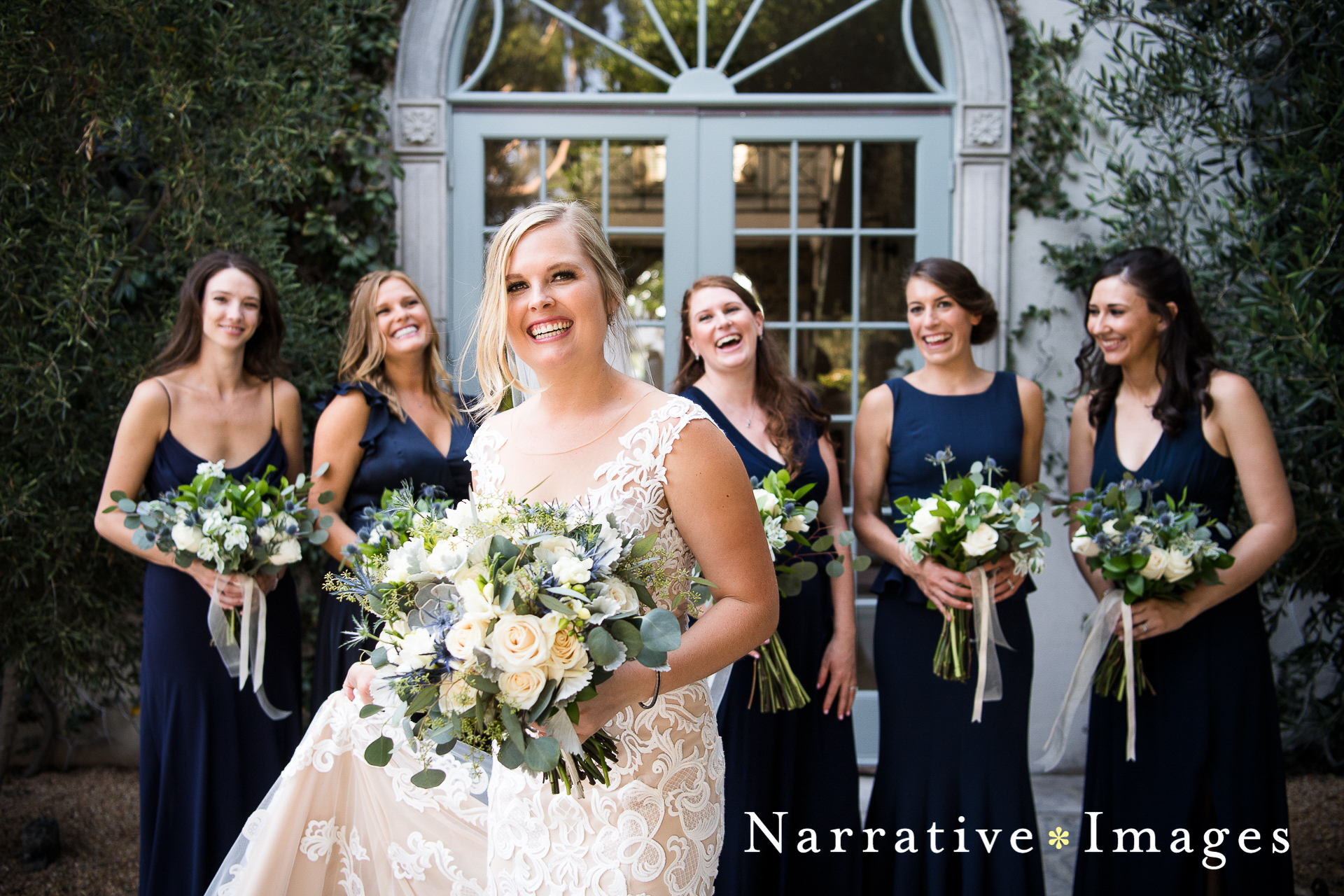 Smiling bride in lace dress and eucalyptus thistle bouquet with her bridesmaids in navy blue dresses
