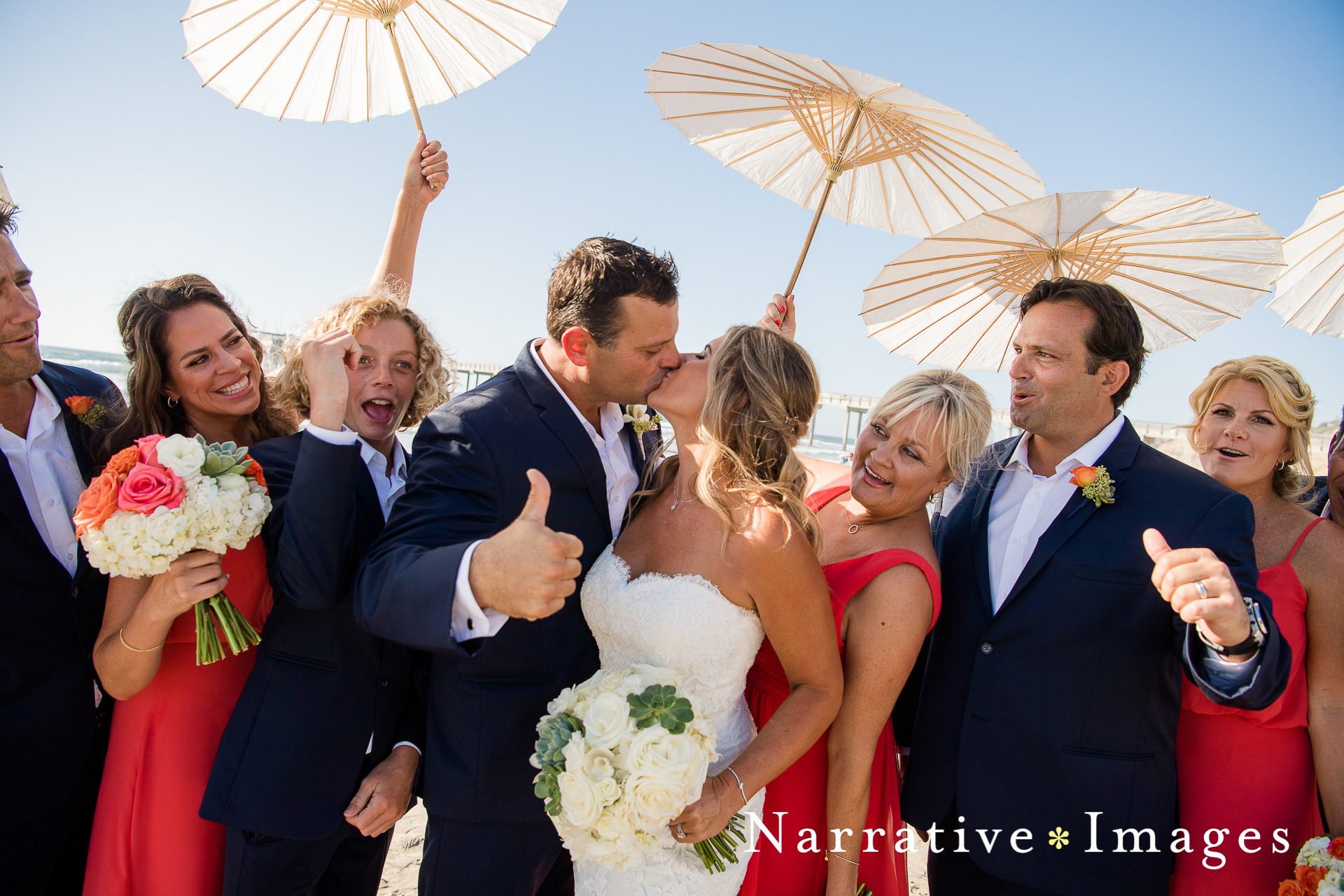 Bride and groom and wedding party kiss with thumbs up on the beach at Scripps Seaside Forum in La Jolla
