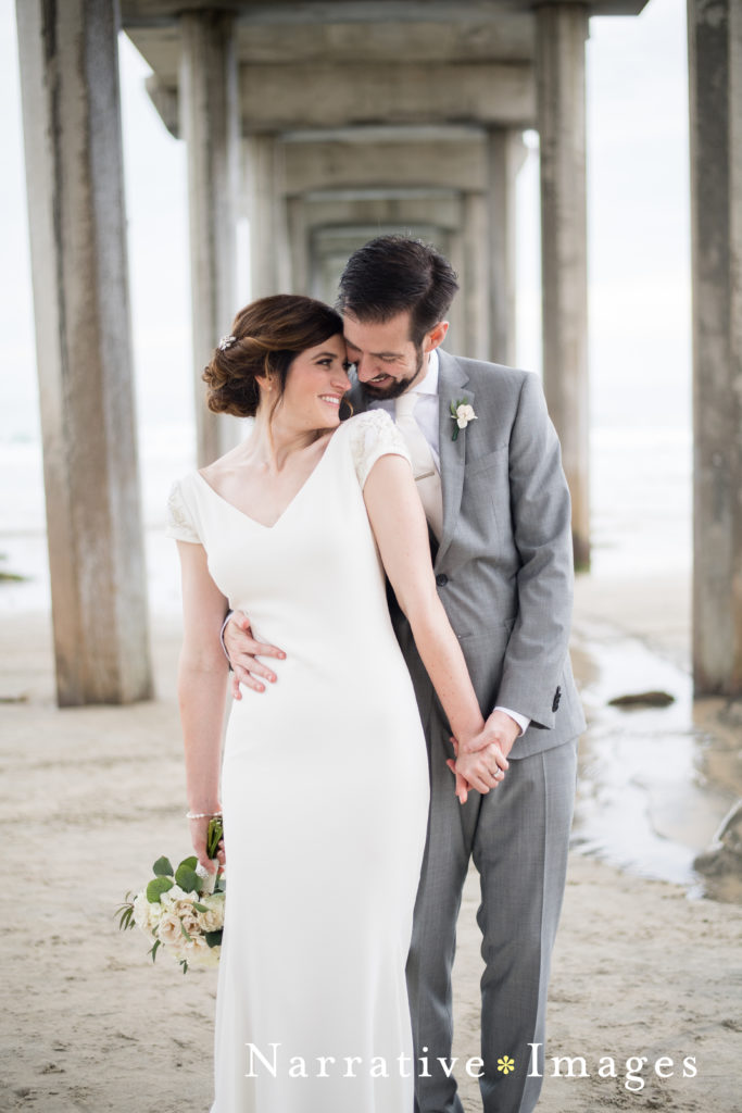 Bride and groom hold hands and snuggle under Scripps Pier in La Jolla, California