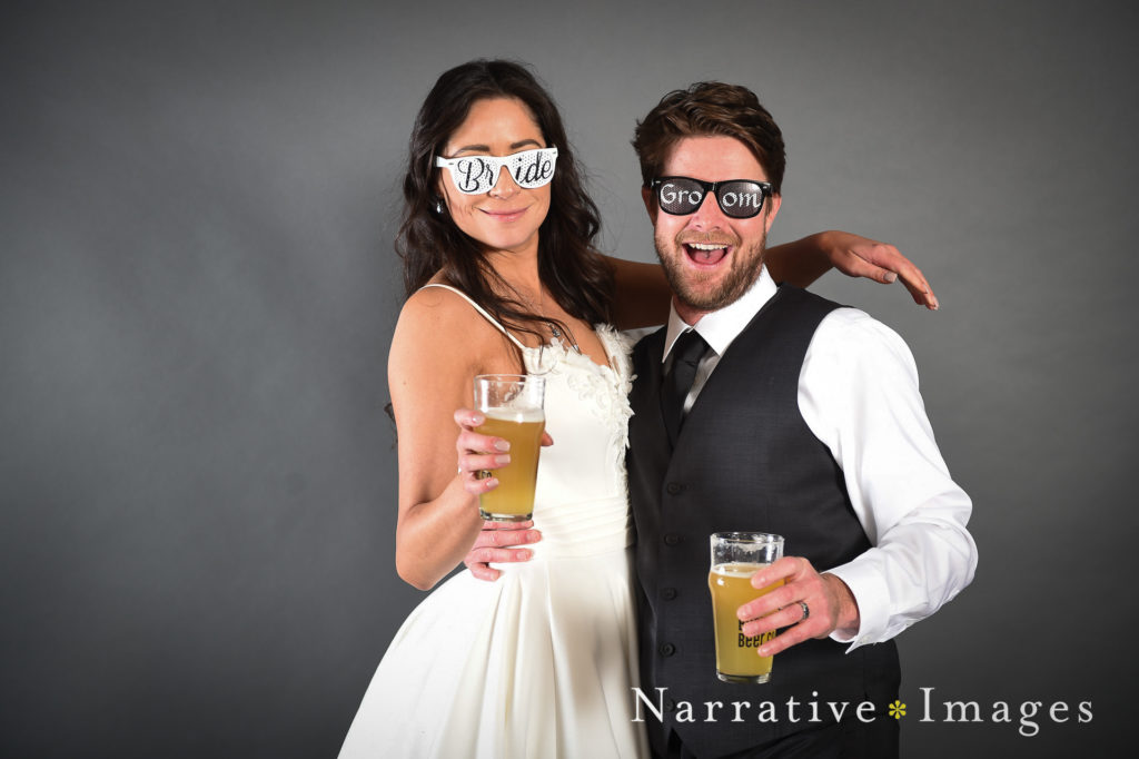Bride and Groom wear shades that say "bride" and "groom" in photo studio booth at a wedding at The Lane wedding venue in San Diego