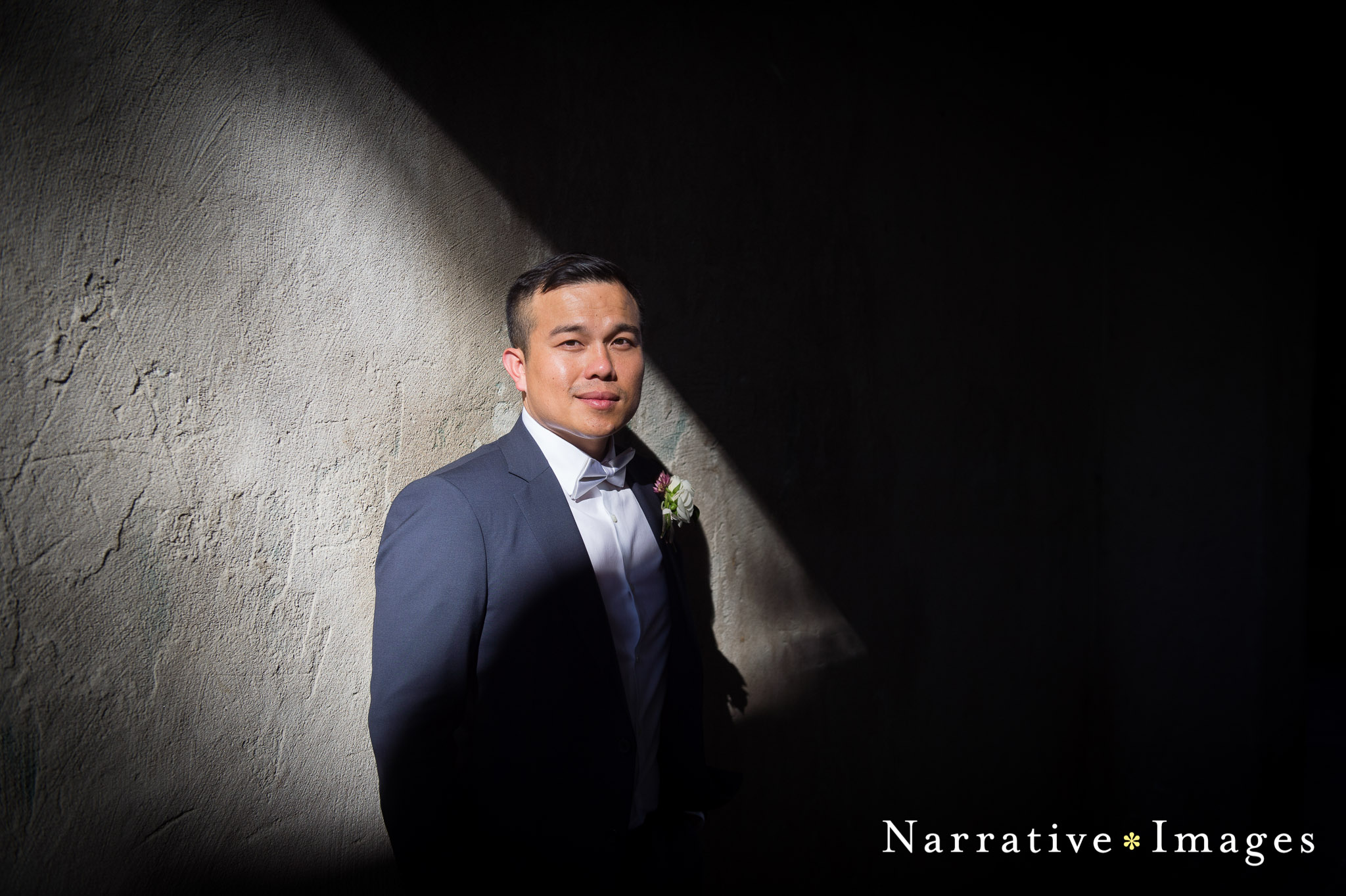 Groom with bowtie stands in light in Balboa Park against concrete wall