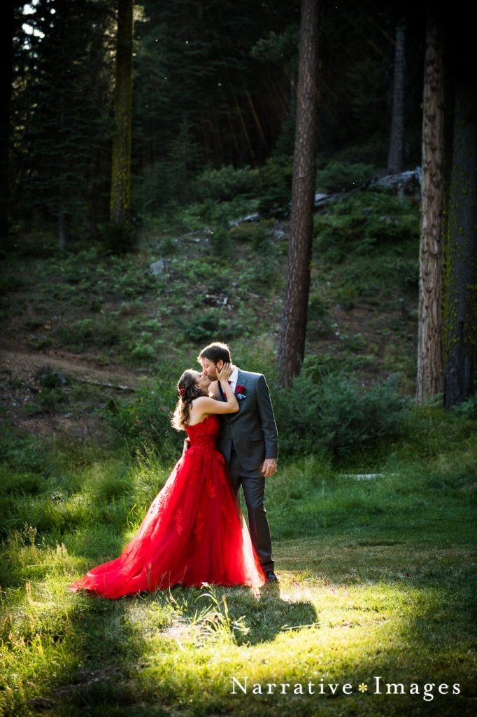 Bride is red wedding dress kisses groom in woods at outdoor wedding surrounded by pine trees