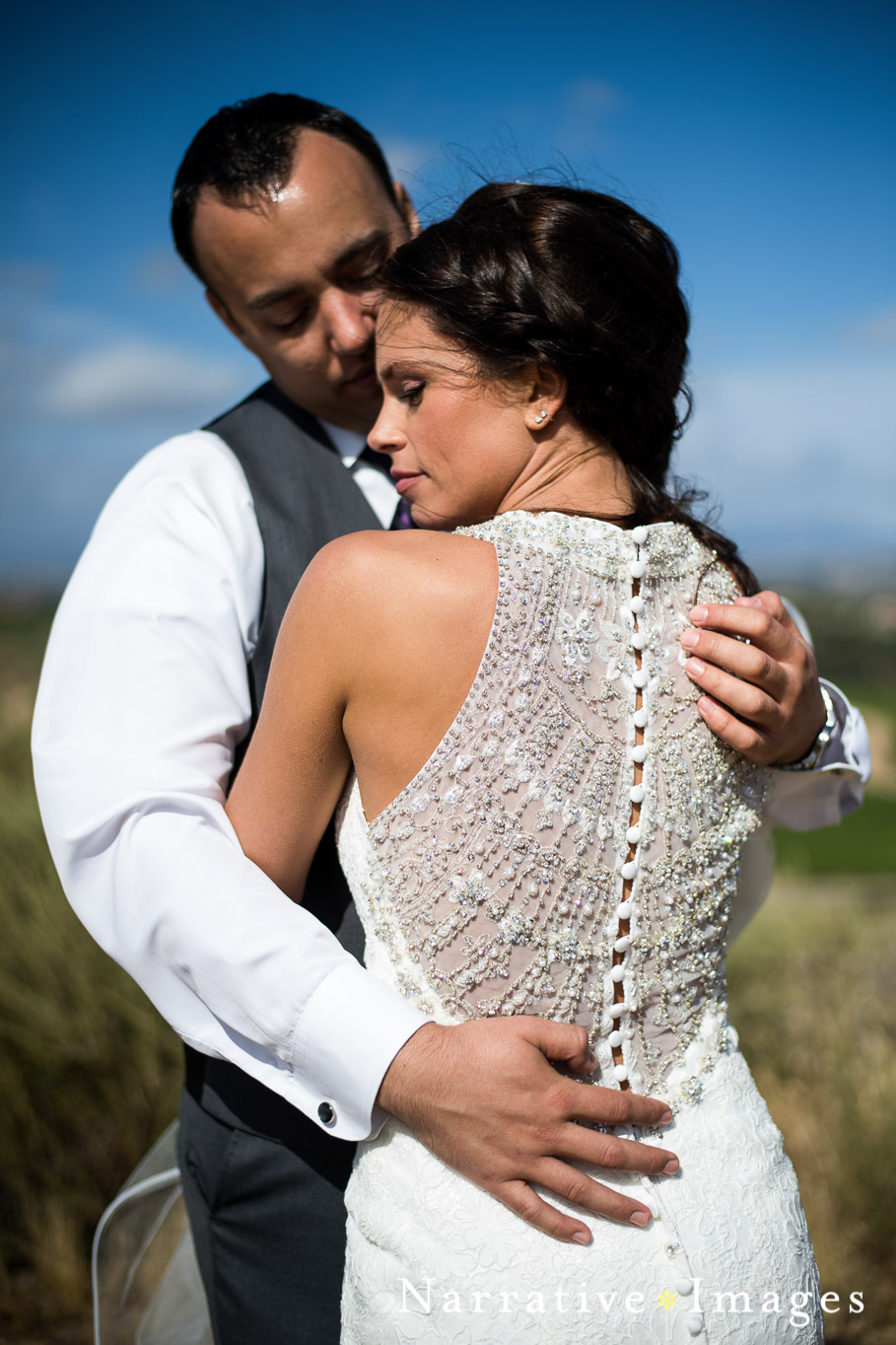 Wedding couple embrace in bright sunlight with halter lace bridal gown that has sheer back with beading 