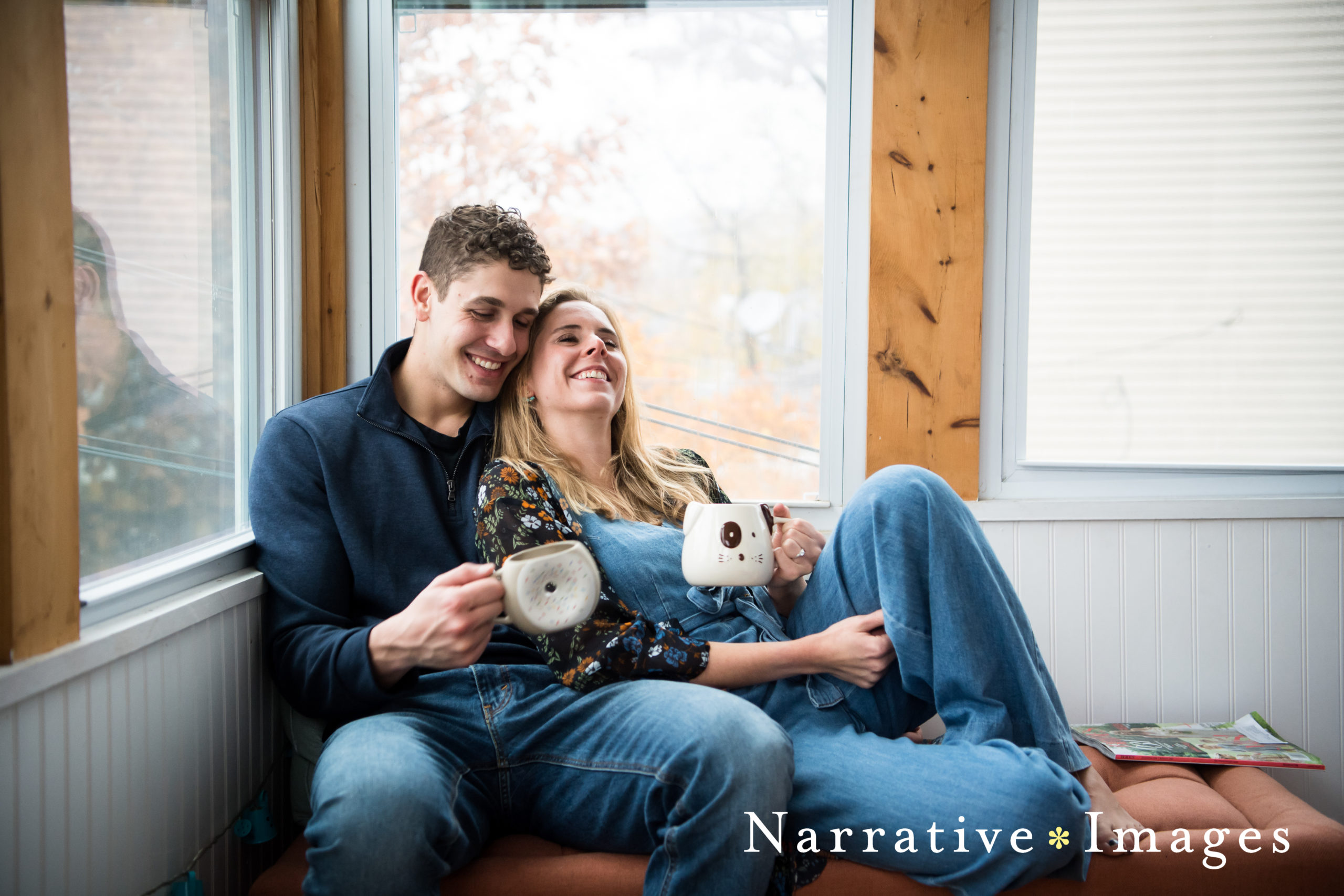 Couple holding mugs and leaning on one another laughing on chaise by windows in their home.