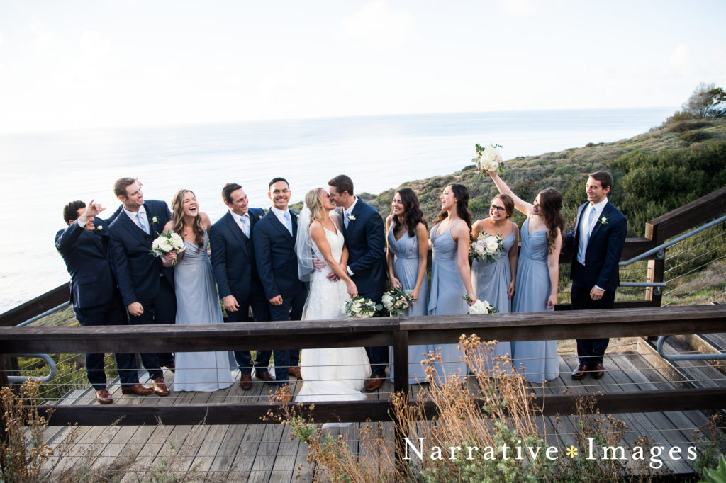 Wedding party cheers as bride and groom kiss on stairs overlooking Scripps Beach in La Jolla, California
