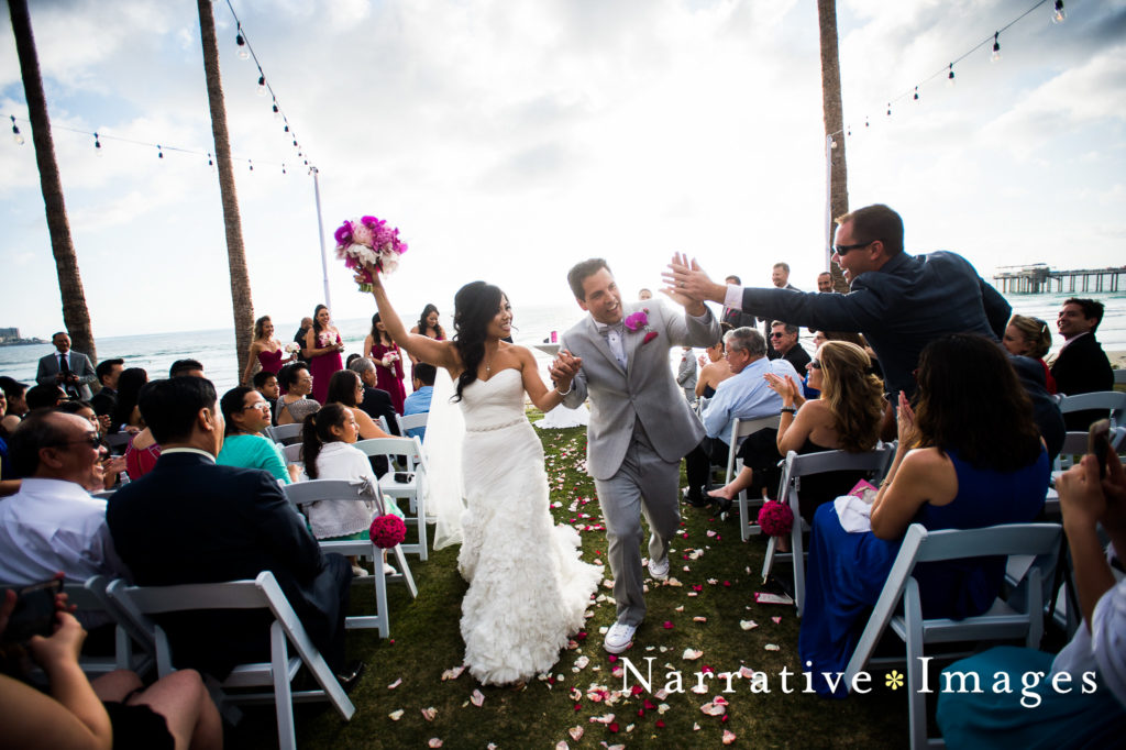 Bride and groom walk down the aisle as groom high fives a guest on the beach at Scripps Seaside Forum